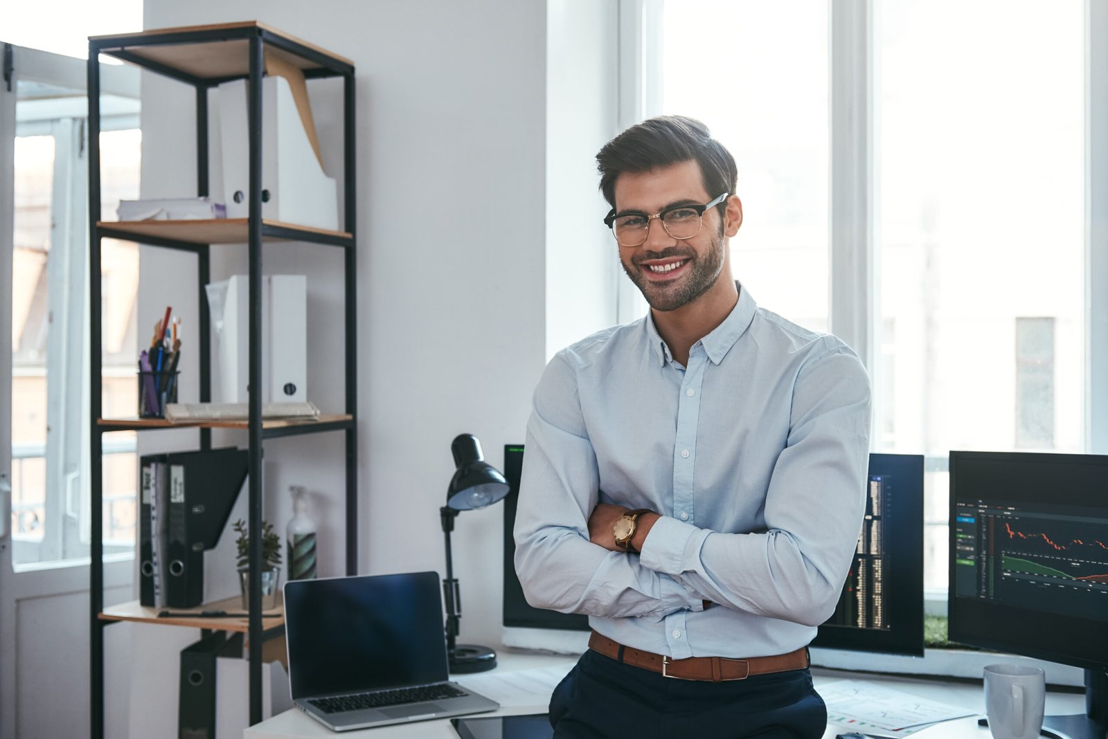 Happy trader. Cheerful businessman in formal clothes and eyeglasses is keeping arms crossed, smiling and looking at camera while standing at office. Stock broker. Forex market. Trade concept